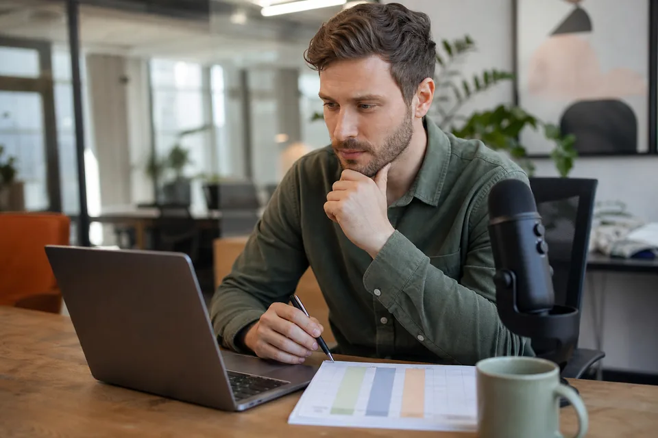 Brand marketing manager at a desk comparing podcast and YouTube sponsorship options on a laptop next to a printed budget sheet