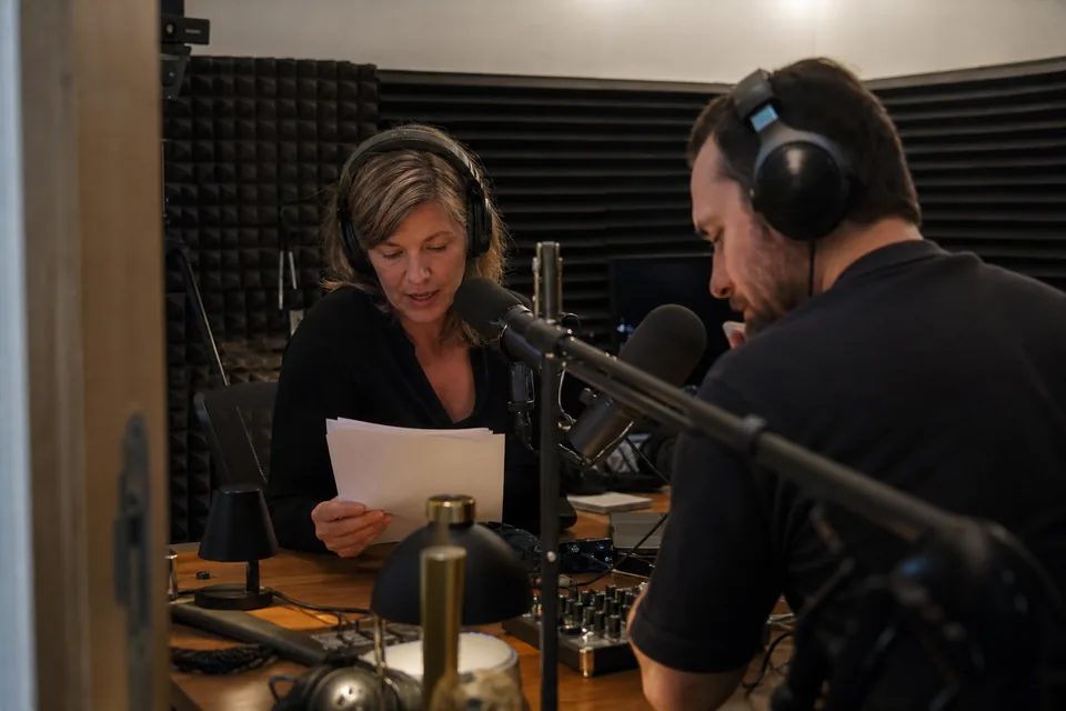 Two podcast hosts at studio microphones recording a host-read ad segment, one reading from a printed sponsor brief